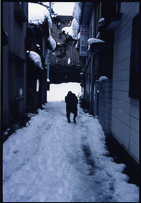 雪が降り積もっている西方寺入口の道を一人の老婆が歩いている写真