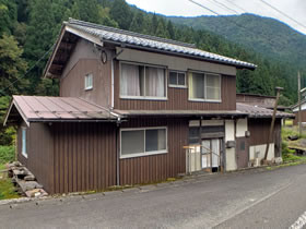 山を背景にした茶色の外観を持つ2階建ての家屋の写真