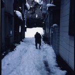 雪が降り積もっている西方寺入口の道を一人の老婆が歩いている写真