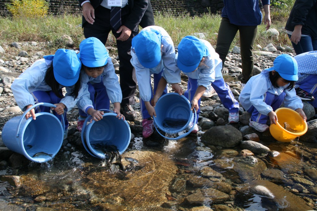 子供たちがバケツに入った稚魚を川に放流している様子の写真